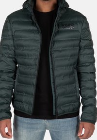 Uomo che indossa una giacca trapuntata verde scuro con il logo "WRONG FRIENDS", una maglietta nera e jeans azzurri chiari, in piedi con le braccia rilassate.