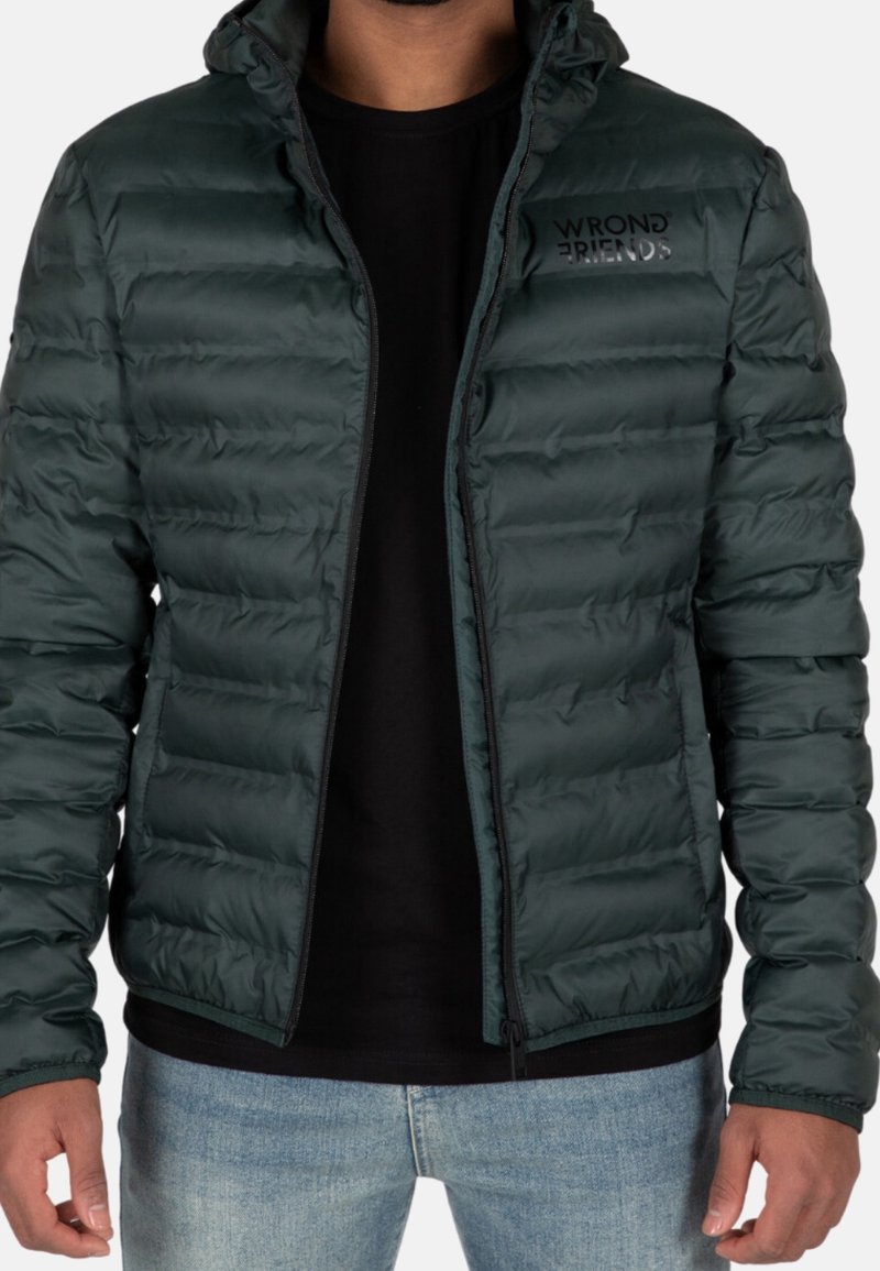 Uomo che indossa una giacca trapuntata verde scuro con il logo "WRONG FRIENDS", una maglietta nera e jeans azzurri chiari, in piedi con le braccia rilassate.