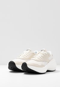 Ljust beige sneakers med en texturerad mesh-överkappa, vita accenter och en tjock svart gummisula. Har en rundad, plattform design.
