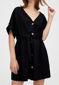 Czarna krótka sukienka z dekoltem w serek, krótkimi rękawami i talią ze sznurkiem; posiada złote guziki i luźny krój.