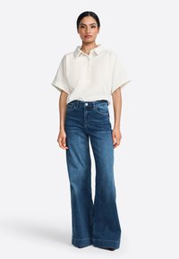 Polo à manches courtes crème avec col et boutons, associé à un jean bleu à jambes larges. Le tissu semble doux ; le jean a une finition soignée et des poches.