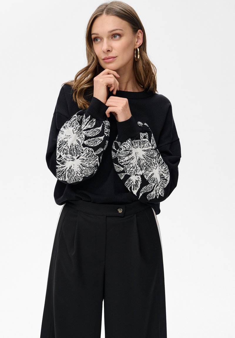 Schwarzer Pullover mit großen weißen Blumenprints an den Ärmeln, Rundhalsausschnitt, lockere Passform, kombiniert mit hoch taillierten schwarzen Hosen.