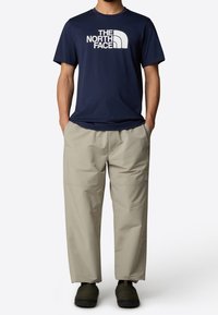 Navy short-sleeve t-shirt featuring a white logo, paired with light beige loose-fitting pants and dark slip-on footwear.