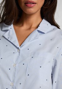 Πουκάμισο με κοντό μανίκι σε ανοιχτό μπλε ριγέ σχέδιο με μικρά σκούρα μπλε μοτίβα καρδιές, κλείσιμο με κουμπιά και μία τσέπη στο στήθος. Λείο ύφασμα.