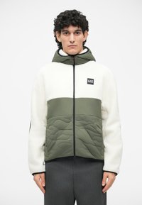 Jachetă cu glugă, având partea superioară din fleece alb și partea inferioară din material matlasat verde. Fermoar negru și patch cu logo pe piept. Țesătură texturată.