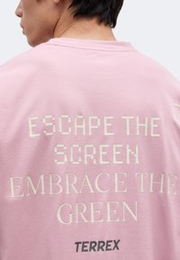 Άτομο που φοράει ροζ μπλούζα με το κείμενο "ESCAPE THE SCREEN EMBRACE THE GREEN" και το λογότυπο "TERREX" στην πλάτη.