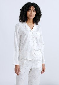 Νεαρή γυναίκα με μπούκλες, ντυμένη με λευκή πυτζάμα μακρυμάνικη με μικρά κόκκινα καρδιές, στέκεται μπροστά σε απλό φόντο.