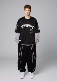 Zwarte oversized t-shirt met witte grafische tekst en illustraties, gelaagd over een grijs lang mouwt-shirt. Losse zwarte broek met witte naden.