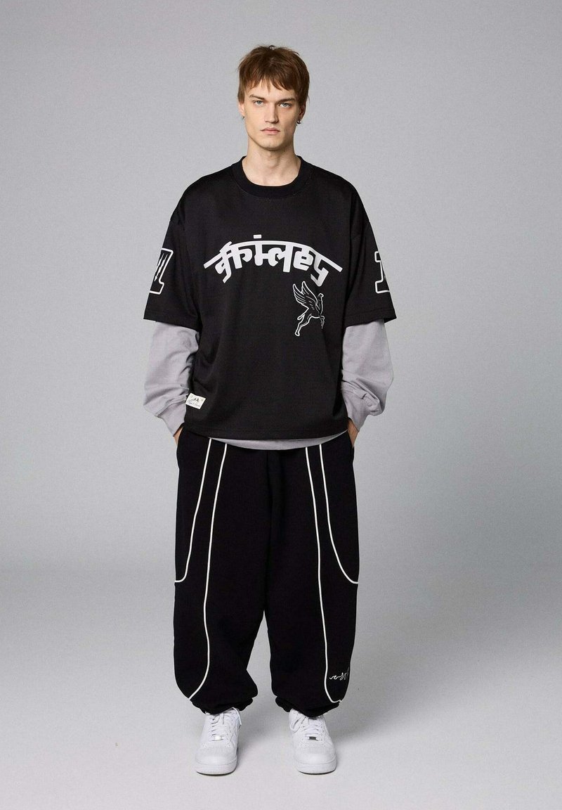 Zwarte oversized t-shirt met witte grafische tekst en illustraties, gelaagd over een grijs lang mouwt-shirt. Losse zwarte broek met witte naden.