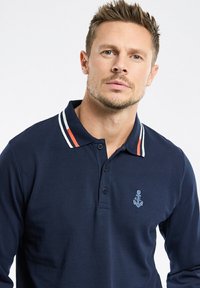 Polo-shirt marine en coton texturé, avec une patte de boutonnage à deux boutons, un col rayé blanc et rouge, et un logo ancre brodé.