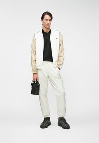 Track jacket και παντελόνα σε λευκό και μπεζ νάιλον, με μαύρο μπλουζάκι από κάτω και μικρή μαύρη τσάντα. Τα υφασμάτινα αθλητικά παπούτσια ολοκληρώνουν το σύνολο.