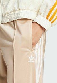 Beige träningsbyxor med sidostrikar, som har en liten vit Adidas-logotyp. Tyget verkar slätt och något glänsande, med en ficka med dragkedja.