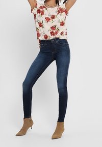 Top a maniche corte con stampa floreale, con fiori rossi su uno sfondo crema, abbinato a jeans skinny blu e stivaletti beige alla caviglia.