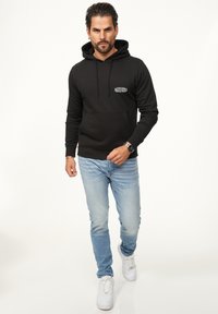 Schwarzer Kapuzenpullover mit einer Fronttasche und Logodetails, kombiniert mit hellblauen Jeans und weißen Sneakers. Glatte Stofftextur.
