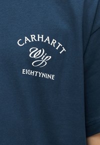 Μπλε ύφασμα με λευκό κείμενο που γράφει "Carhartt Wip Eightynine" σε μικτούς χαρακτήρες σε μανίκι ρούχου.