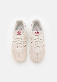 Witte sneakers met een suède textuur, rode strepen en geperforeerde neus. Voorzien van een logo op de tong en een gewatteerde binnenzool. Klassiek laag model.