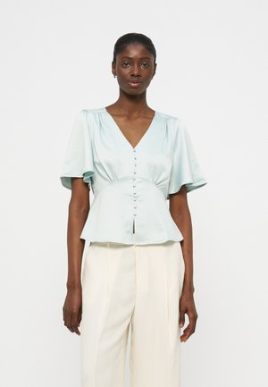 Žena v svetlomodrej krátkorukávovej blúzke na gombíky s výstrihom do V a bielych širokých nohaviciach s vysokým pásom, stojaca pred bielym pozadím.