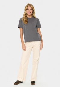 Grå t-shirt med kort ärm och subtila dekorationer, tillsammans med ljusbeige vida byxor och mörka skor, stående mot en enfärgad bakgrund.