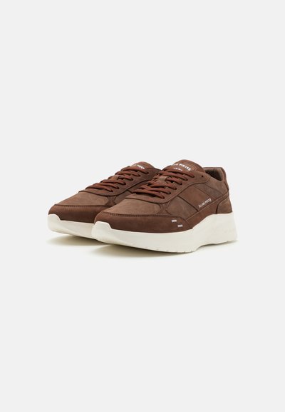 Pár hnědých semišových tenisek s silnými bílými podrážkami a tkaničkami, nakloněné vlevo na bílém pozadí, značkované "Filling Pieces" na jazyku a boku.