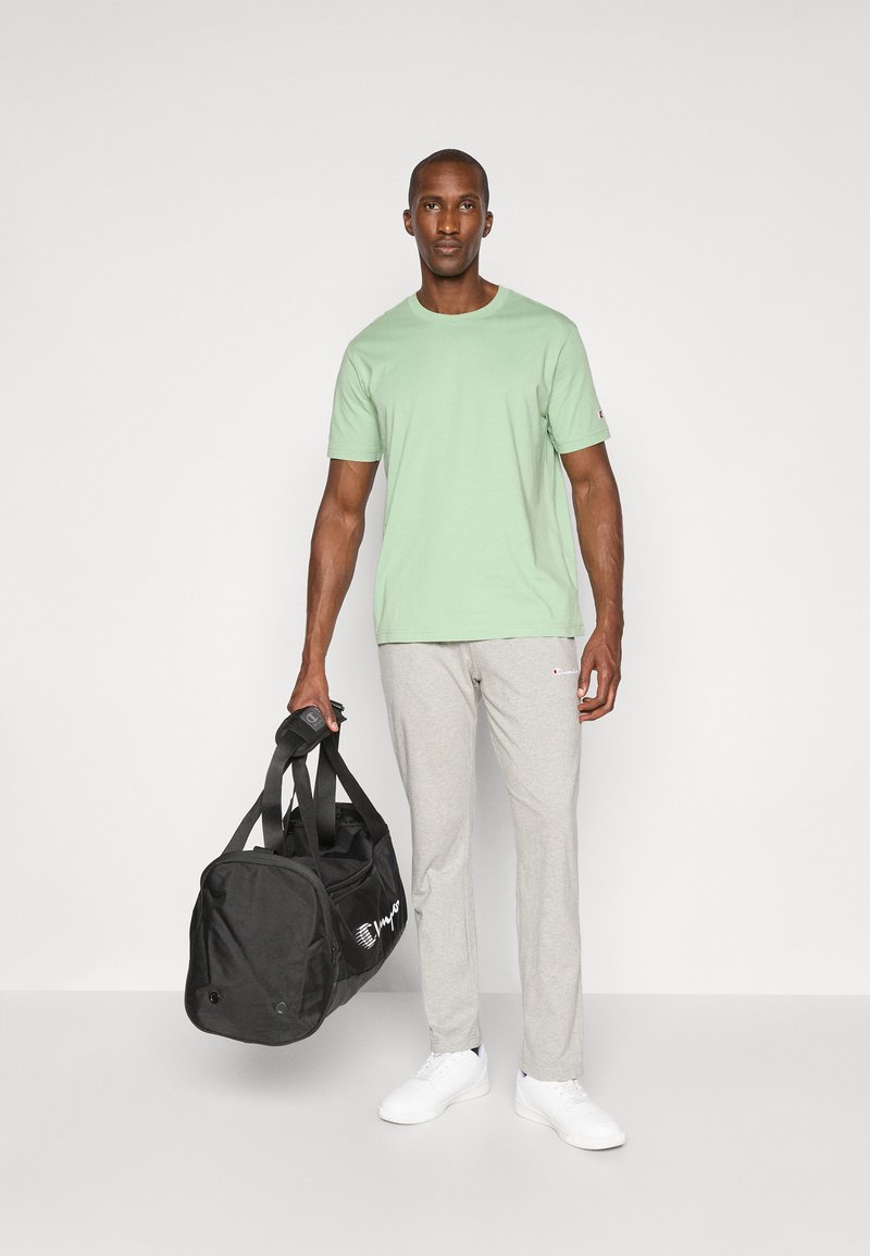 Grünes Baumwoll-T-Shirt, graue Jogginghose und weiße Sneakers. Schwarze Sporttasche mit Logo. Modell steht vor einem einfarbigen Hintergrund.