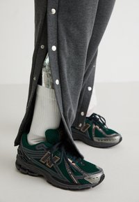 Graue Sporthosen mit Druckknopfverschluss an den Seiten, die ein reflektierendes silbernes Innenfutter zeigen. Kombiniert mit grünen Mesh-Sneakern von New Balance und weißen Socken.