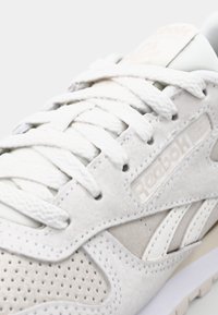 Light gray suede sneaker with white accents, featuring a textured upper, perforations, and flat laces. Reebok logo on the side.