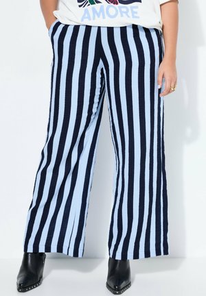 Henkilö, jolla on yllä vaaleansiniset ja mustat pystysuorat raidalliset leveälahkeiset housut, mustat nilkkurit ja valkoinen t-paita, jossa lukee "AMORE".