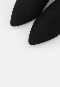 Mocasines negros de ante con punta afilada, que presentan una textura suave y un diseño minimalista, con una parte delantera cónica y sin marcas ni adornos visibles.