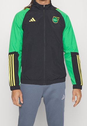 Persona che indossa una giacca con zip nera e verde con strisce gialle Adidas e un logo della squadra di calcio della Giamaica sul petto.