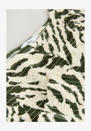 Zelená a krémová vzorovaná tkanina s marsknutou texturou, zdobená listovým motivem. U štítku nahoře je nápis „NEXT“.