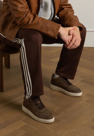 Bruine leren sneakers met een gestructureerde afwerking, donkere veters en een witte zool. Gecombineerd met bruine broek met witte strepen.