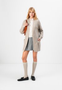 Frau in beigem Cardigan, hellrosa Bluse, grauem Faltenrock, knielangen beigen Socken, schwarzen Loafers, die eine braune Schultertasche hält und vor einem weißen Hintergrund steht.