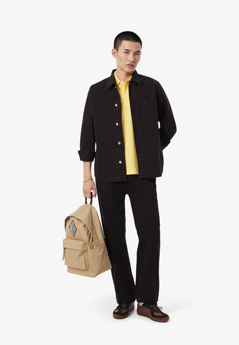 Giacca di jeans nera sopra una polo gialla, abbinata a pantaloni neri e sneakers marroni. Zaino beige con toppe logo e tasca frontale.