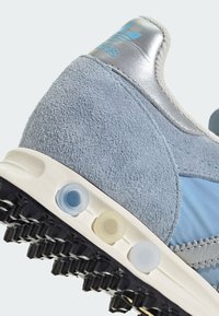 Ανοιχτό μπλε αθλητικό παπούτσι με επάνω μέρος από σουέντ και ύφασμα, ασημένιες λεπτομέρειες, μαύρη ελαστική σόλα με καρφιά και τρεις στρογγυλές πρίζες σε διαφορετικά χρώματα.