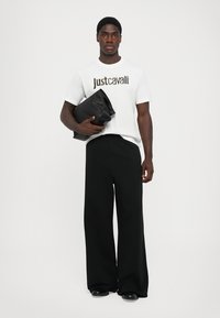Balts T-krekls ar "Just Cavalli" logo melnā un metāliskās krāsās, saskaņots ar platām melnām biksēm un melnu clutš somu.