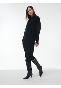 Schwarzer Strickhoodie mit durchgehendem Reißverschluss, zwei Fronttaschen und einer lockeren Passform, kombiniert mit einem figurbetonten schwarzen Rock und knielangen schwarzen Stiefeln.