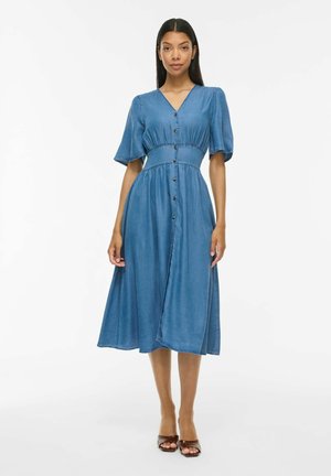 Kvinde står i en blå denim-midi-kjole med knapper foran, korte ærmer og taljeret snit samt brune sandaler mod en enkel hvid baggrund.