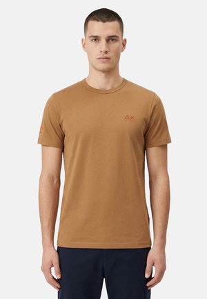 Uomo in piedi di fronte, indossa una maglietta marrone semplice a maniche corte con un piccolo logo sul petto e pantaloni scuri su uno sfondo chiaro.