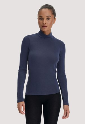 Junge Frau mit zurückgebundenen Haaren, die einen marineblauen Langarm-Rollkragenpullover und schwarze Hose vor hellem, einfarbigem Hintergrund trägt.