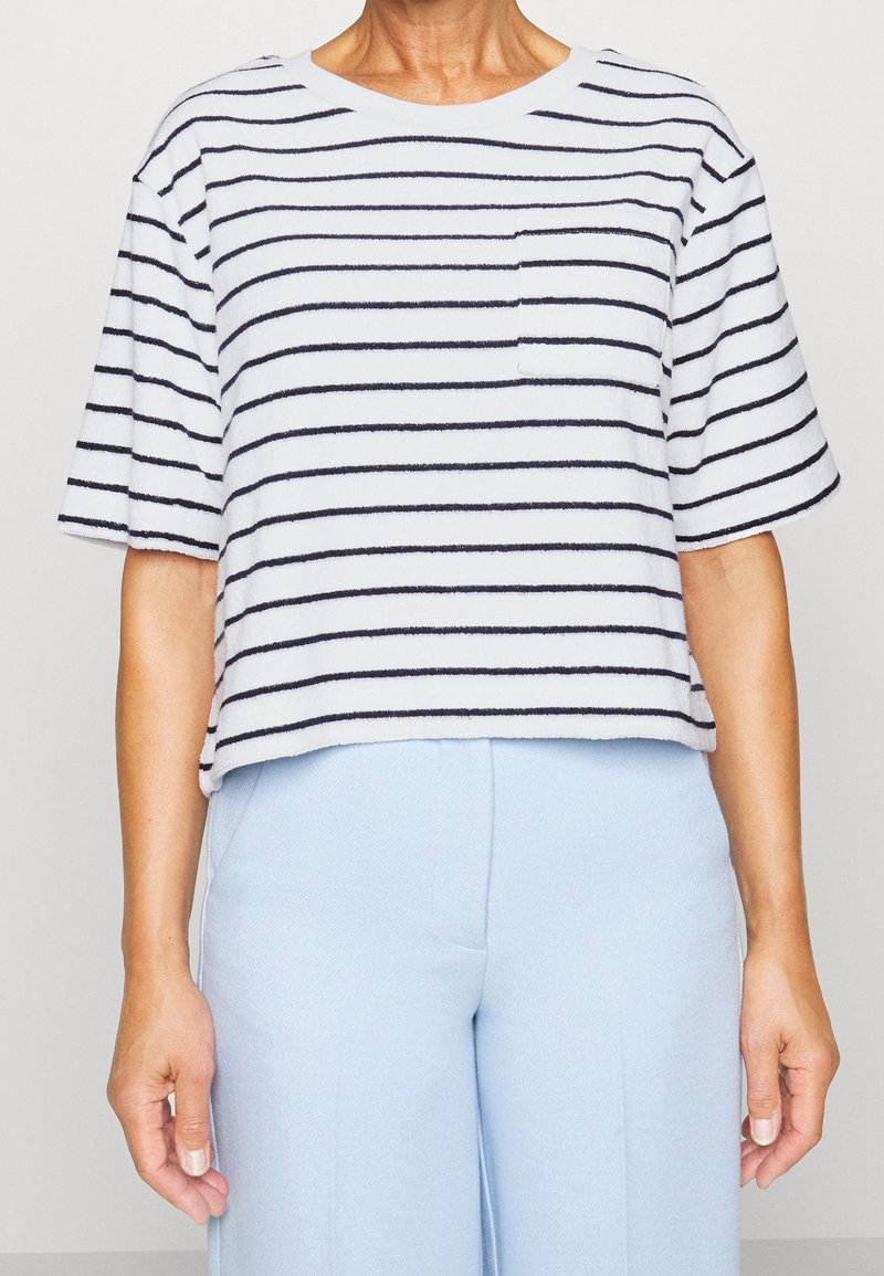 Femme portant une chemise blanche à manches courtes avec des rayures horizontales noires et un pantalon taille haute bleu clair.