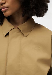 Nahaufnahme einer Person, die eine beige North Face-Jacke mit spitzem Kragen trägt, auf der das geprägte Markenlogo auf der Brust zu sehen ist.