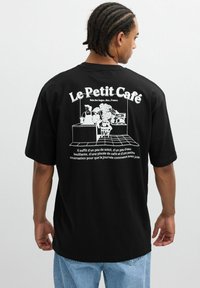 Hombre con camiseta negra de gran tamaño con un gráfico blanco de una escena de café, plantas y texto en francés "Le Petit Café" en la parte trasera.