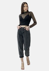 Schwarzes, langärmeliges Cropped-Top aus Netz mit V-Ausschnitt, kombiniert mit hoch taillierten dunkelgrauen Jeans und weißen Riemchen-Heels. Das Model posiert mit den Händen in den Taschen.