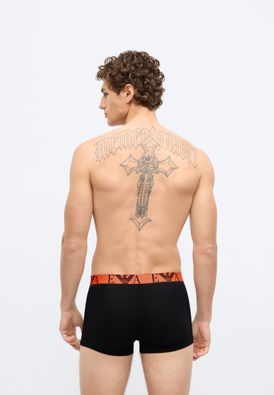 Boxer neri con una fascia in vita arancione che mostra il logo del marchio. Il modello ha un tatuaggio di una spada e del testo sulla schiena.