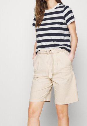 Kobieta ubrana w krótki rękaw z czarno-białymi paskami, włożona w beżowe bermudy do kolan z paskiem, ręce w kieszeniach.