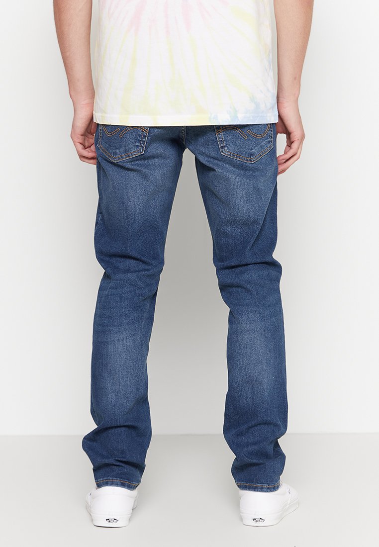 Jeans de denim azul claro con corte recto, costuras en contraste y dos bolsillos traseros. Llevados con una camisa de tie-dye colorida y zapatillas blancas.