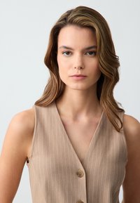 Débardeur beige avec de subtiles rayures verticales blanches, décolleté en V profond, sans manches, et deux boutons marron à l'avant. Texture lisse.