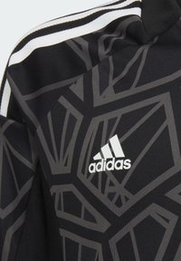 Schwarze Jacke mit einem geometrischen grauen Muster, Stoffstruktur und weißen Akzenten. Das Adidas-Logo ist in weißer Schrift deutlich sichtbar.
