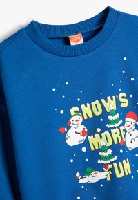 Blå sweatshirt med snögubbe-motiv, julgranar och snöflingor, med ribbad krage och mjuk textur. Texten lyder "SNÖN ÄR MER KUL."