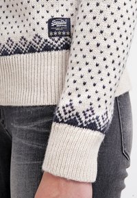 Gros plan sur une manche de pull en maille crème avec motifs noirs et violets et une étiquette de marque SuperDry cousue au-dessus du poignet.